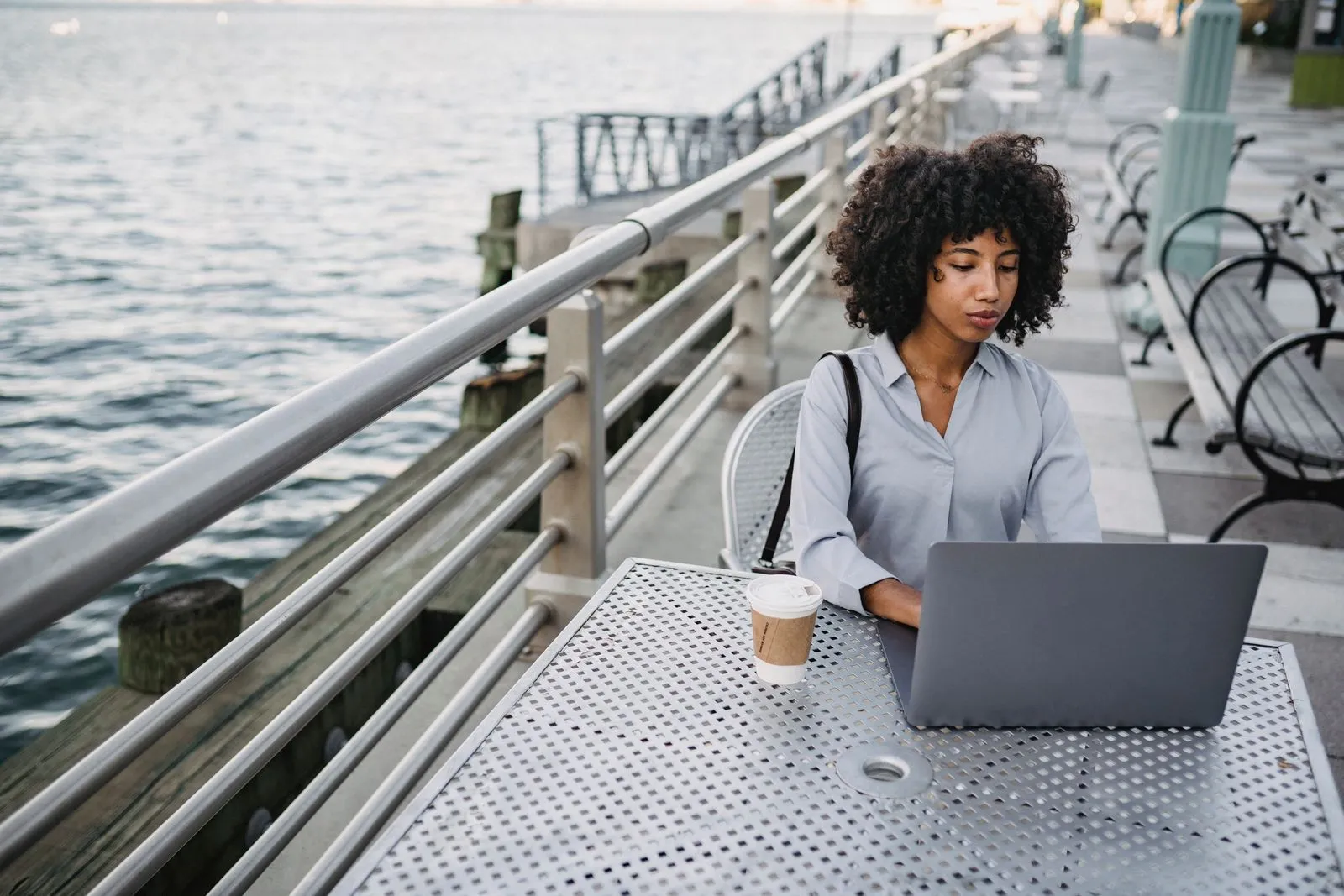 An afro-haired woman working on her laptop at a waterside cafe, with a coffee.
